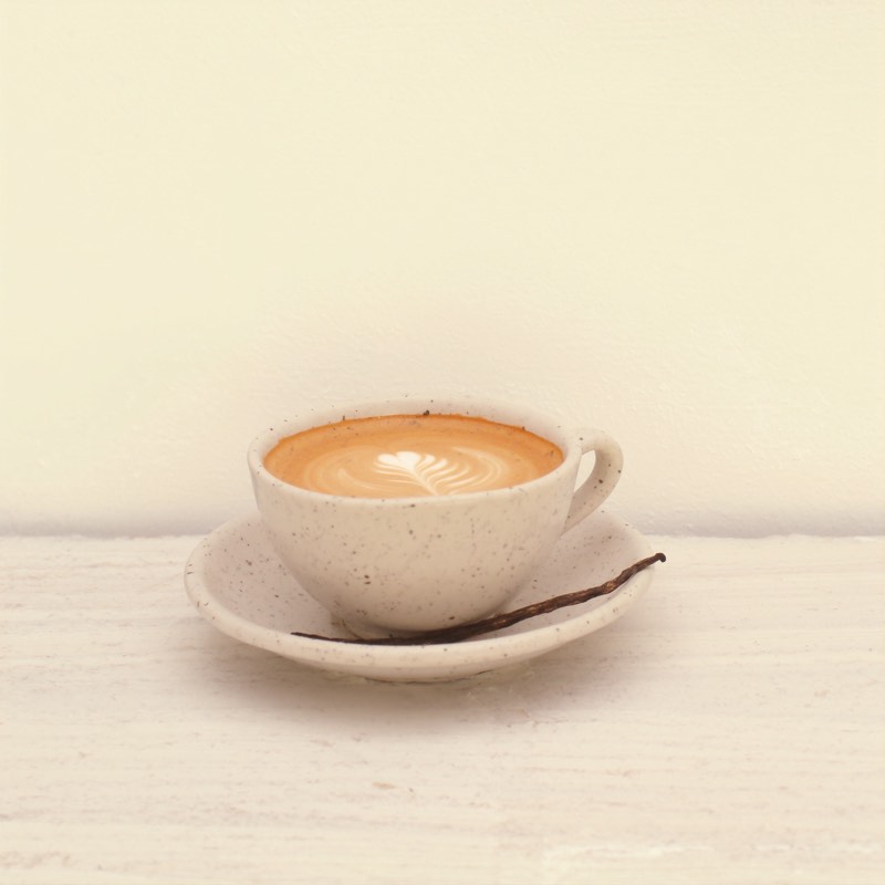 A vanilla latte with feathered latte art and a vanilla pod on the saucer